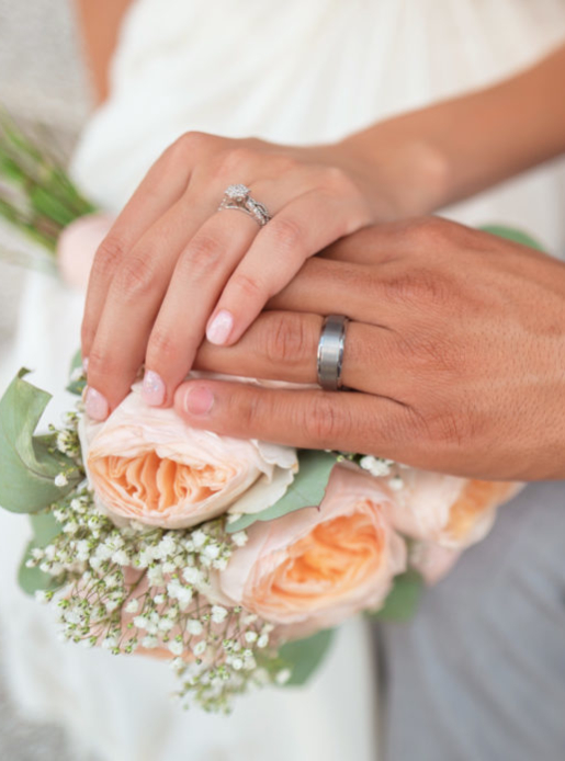 Newlyweds join hands over a bouquet, both wearing their rings