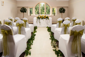 The Park Avenue Room set up for a wedding ceremony, white chairs with green ribbons, ivy down the aisle and sunflower centrepiece on top table
