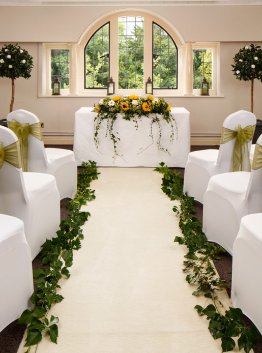 The Park Avenue Room set up for a wedding ceremony, white chairs with green ribbons, ivy down the aisle and sunflower centrepiece on top table