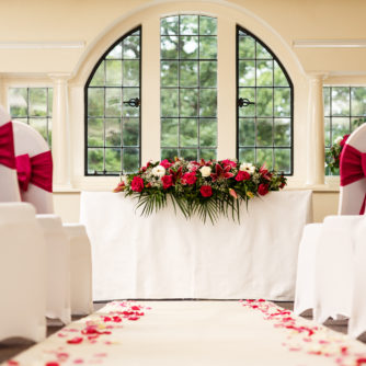 Close up view of The Park Avenue Room set up for a wedding ceremony, white aisle, white chairs, red ribbons and large red and green flower centrepiece on top table