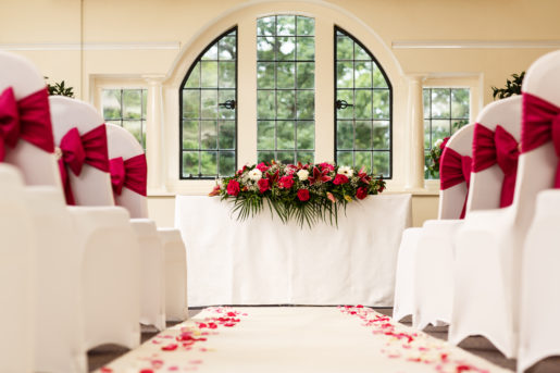 Close up view of The Park Avenue Room set up for a wedding ceremony, white aisle, white chairs, red ribbons and large red and green flower centrepiece on top table